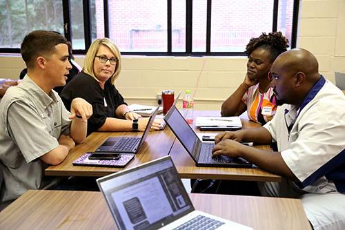 A group of students converse at a table filled with laptops and notebooks